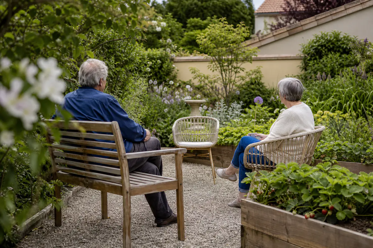 Deux personnes âgées échangent près de bacs d’aromatiques et d’un banc ombragé dans un jardin français fleuri.