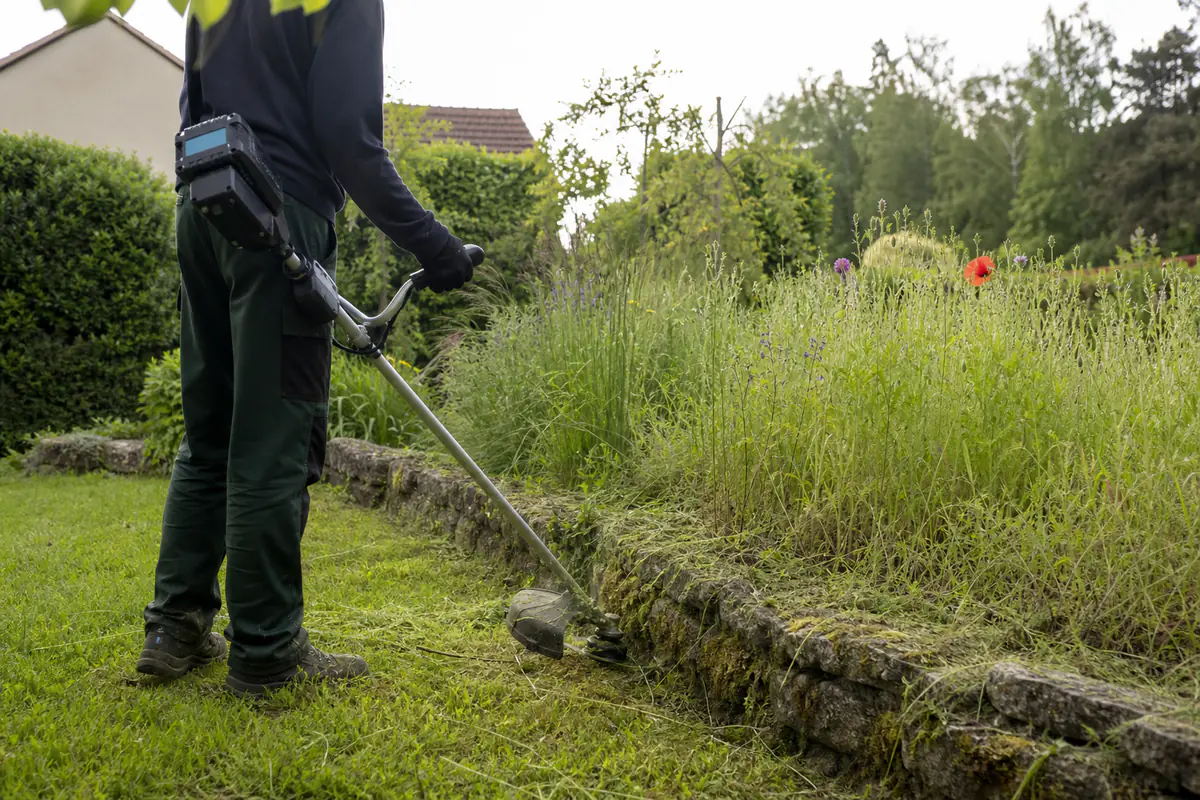 Jardinier équipé d’une débroussailleuse légère coupant des herbes hautes près d’une bordure de jardin