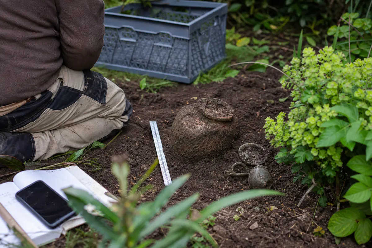 Jardinier français agenouillé devant une jarre ancienne à peine visible dans la terre d’un massif, zone protégée