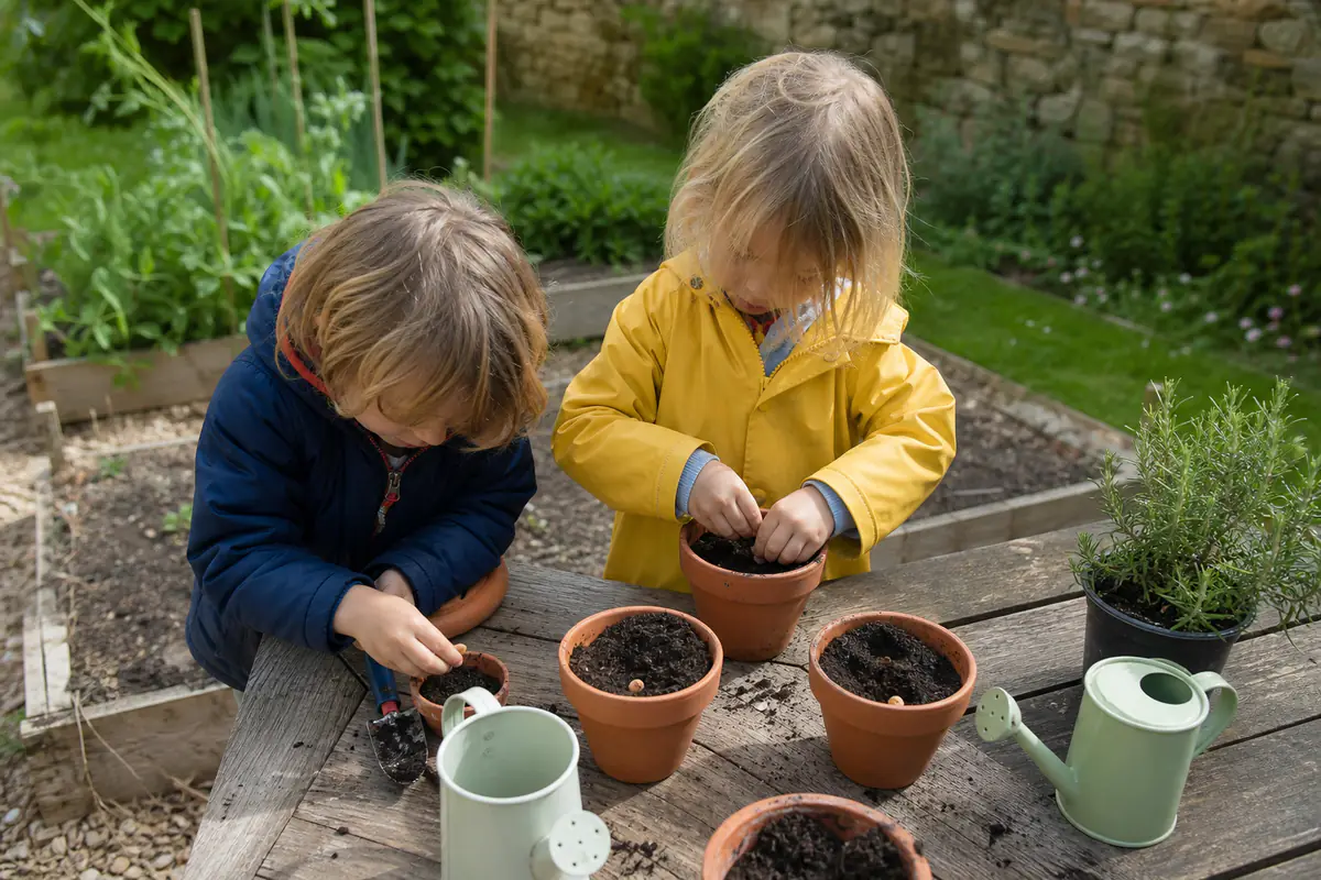 Jeunes enfants semant des graines dans des pots sur une table de jardin, accompagnés d’un adulte attentif