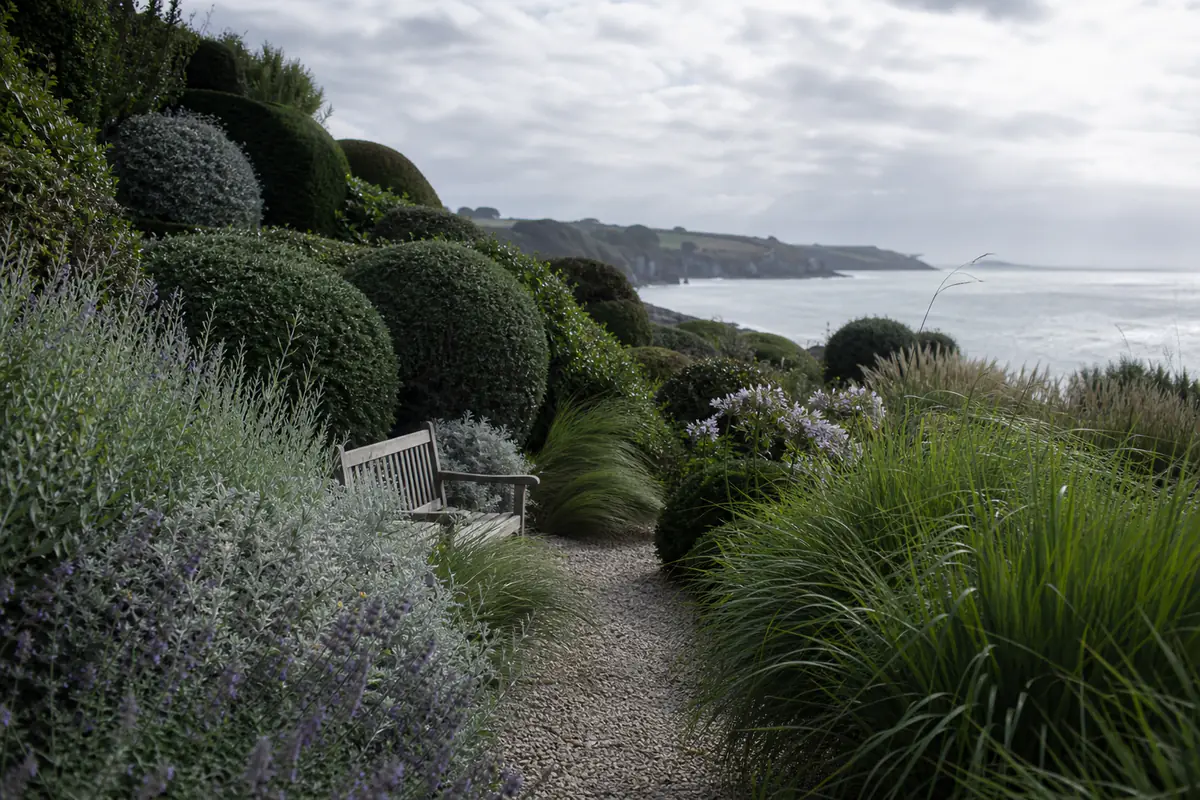 Jardin côtier normand en terrasses, topiaires arrondies et graminées face à la mer sous une lumière claire