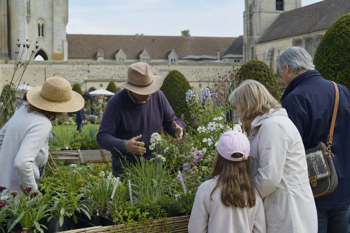 Visiteurs observant des vivaces fleuries et échangeant avec un pépiniériste dans les jardins d’une abbaye