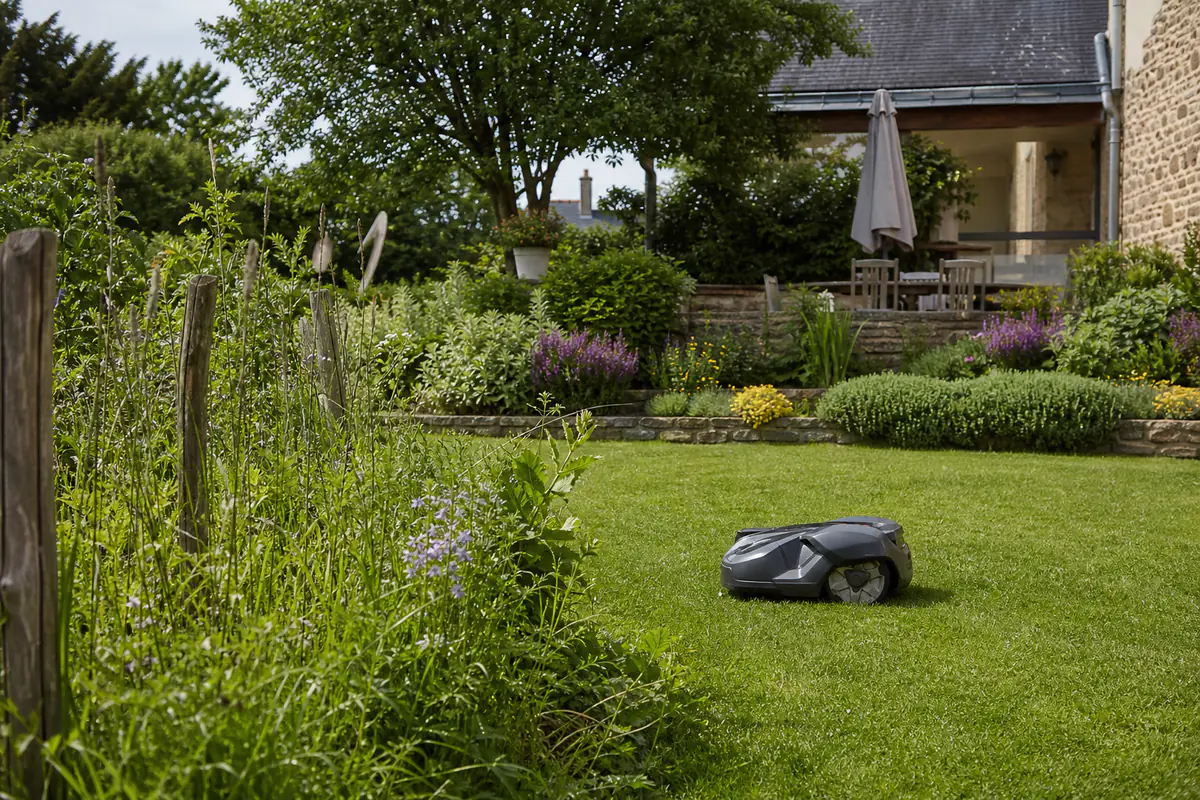 Robot tondeuse compact en marche sur une pelouse familiale bordée de massifs fleuris et d’une terrasse.