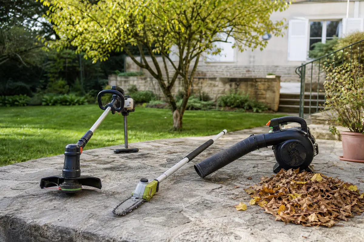 Trois outils de jardin posés sur une terrasse près de bordures, d’un arbre et d’un tas de feuilles sèches.