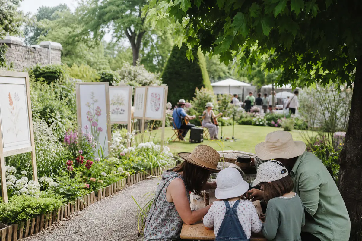Concert acoustique et atelier de semis dans un jardin fleuri, avec visiteurs installés à l’ombre des arbres
