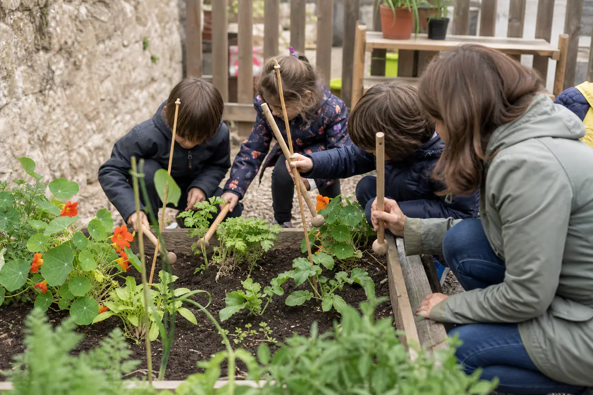 Enfants accompagnés observant des plantes et jouant doucement dans un jardin sonore aménagé dans une cour d’école.