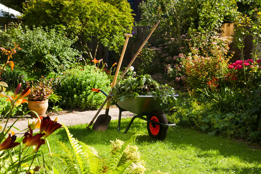 Jardin familial français paillé avec massifs fleuris, arrosoir au pied des plantations et outils manuels posés près d’une allée