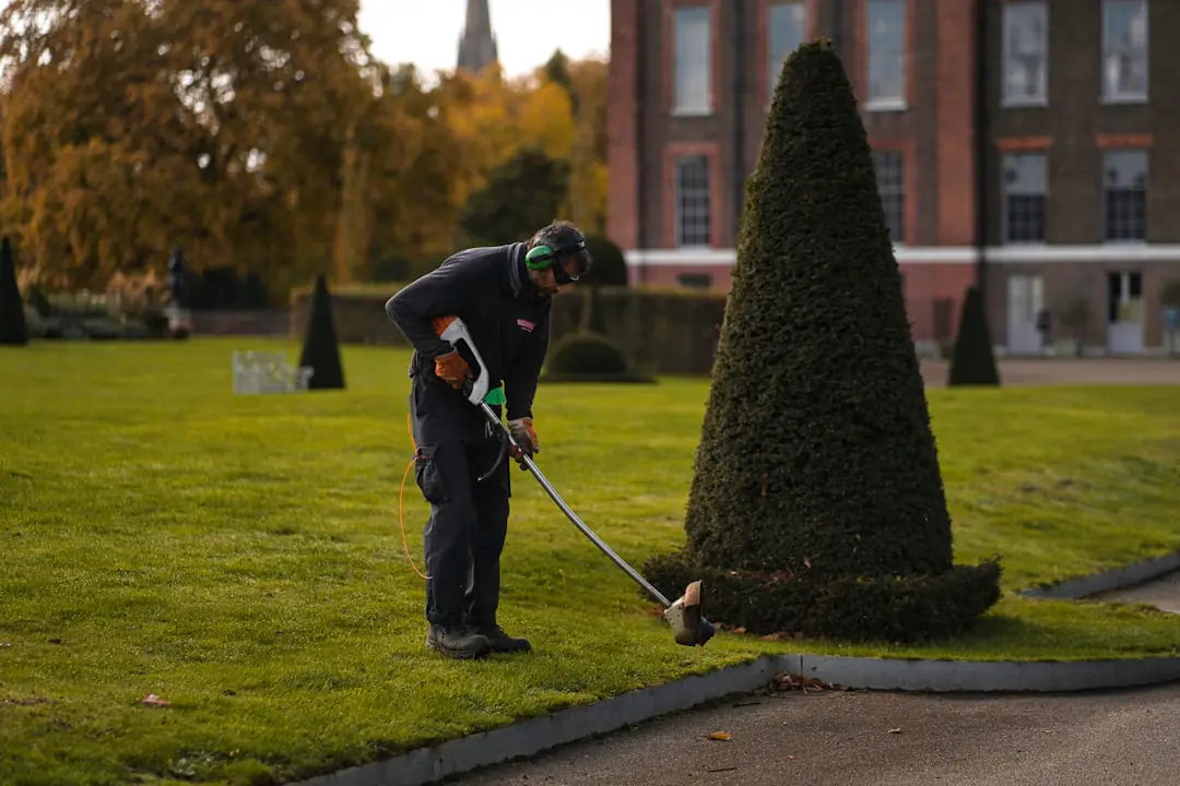 Jardinier utilisant un souffleur sans fil sur une allée bordée de feuilles d’automne, près d’un jardin français