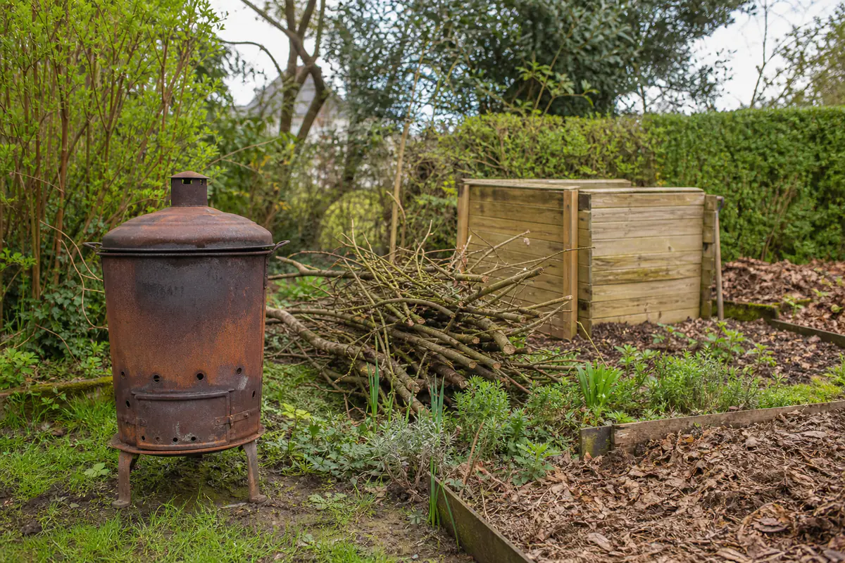 Ancien incinérateur métallique inutilisé près d’un tas de branches et d’un composteur dans un jardin français