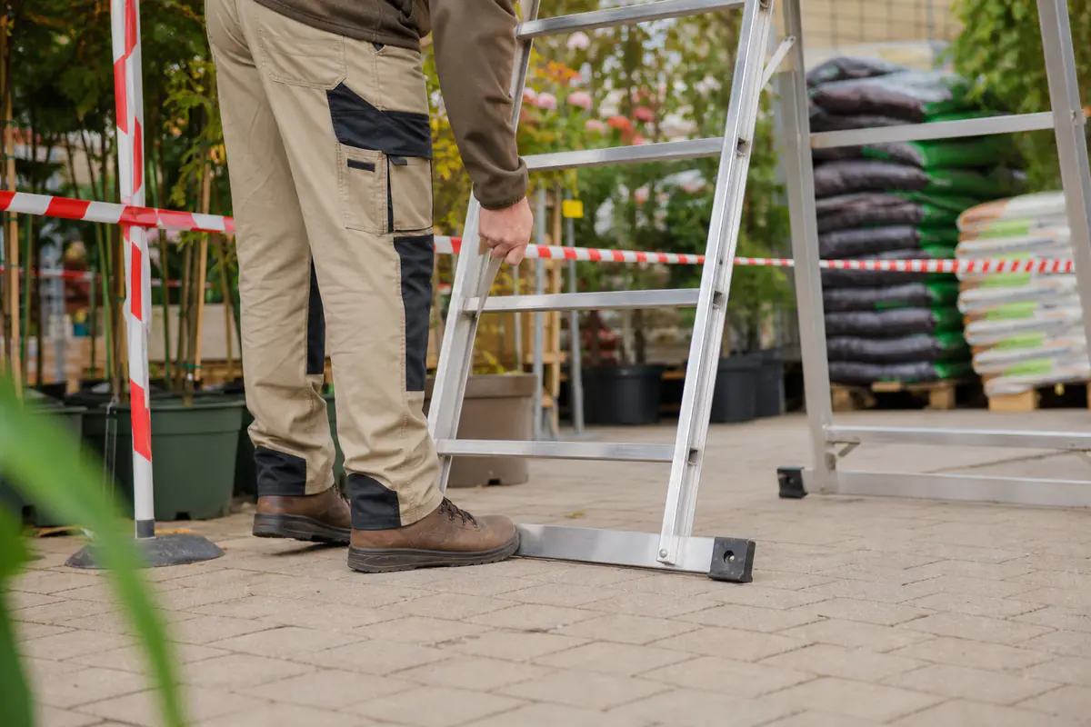 Employé de jardinerie vérifiant l’appui et le stabilisateur d’une échelle sur un sol sec avant utilisation