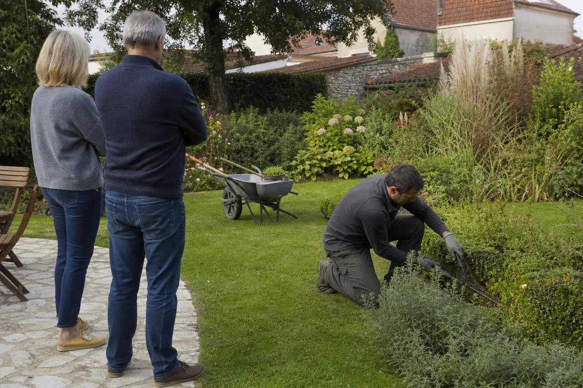 Jardinier entretenant une haie dans un jardin de maison française, tandis qu’un couple observe depuis la terrasse